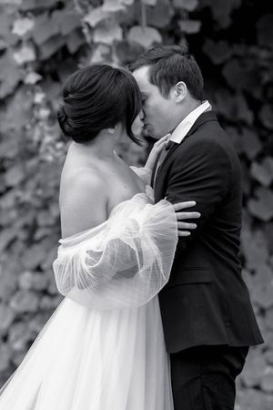 Black and white photo of a bride and groom sharing a tender kiss outdoors against ivy-covered wall.