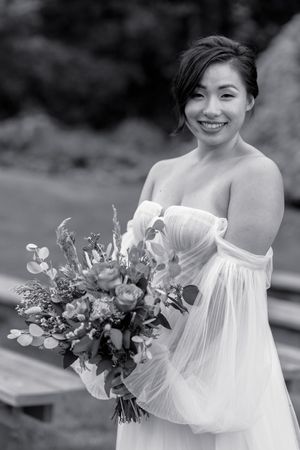 Black and white portrait of a smiling bride holding a lush floral bouquet in an off-shoulder gown.