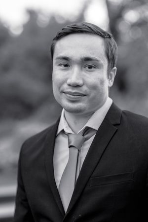 Black and white portrait of a young groom in a dark suit and tie smiling outdoors.