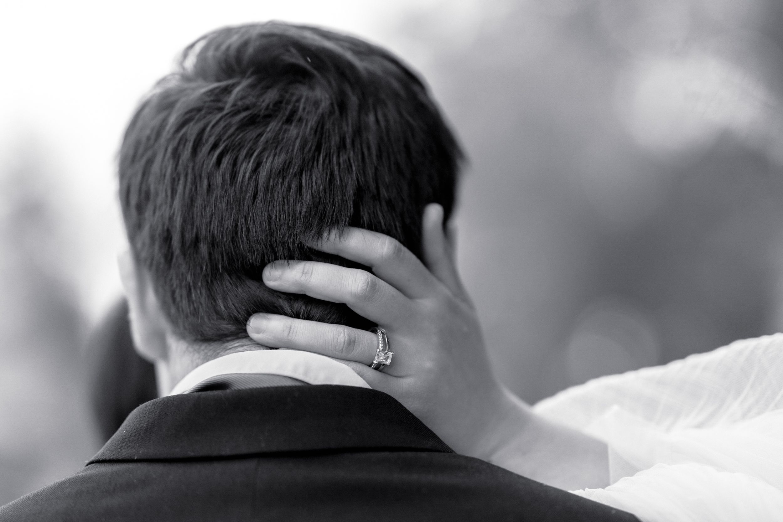 Black and white wedding photo of bride placing hand on grooms neck, diamond ring visible, romantic couple embrace.