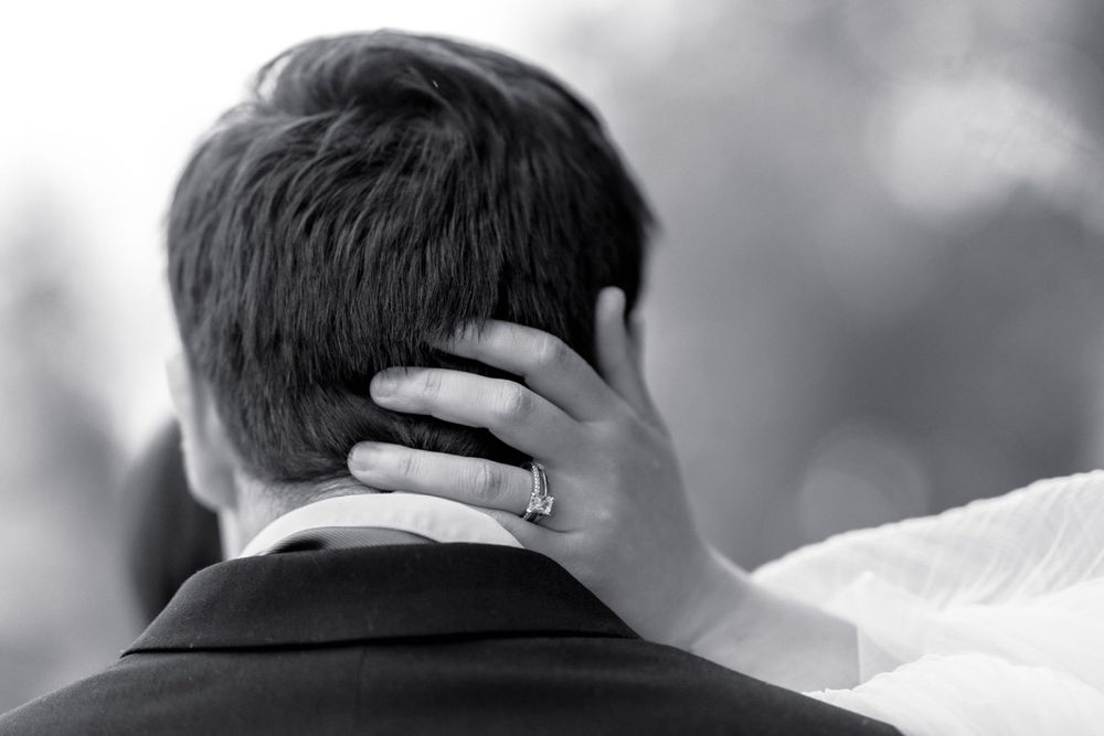 Black and white wedding photo of bride placing hand on grooms neck, diamond ring visible, romantic couple embrace.
