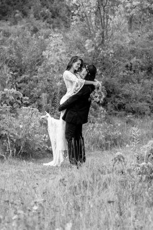 Black and white photo of groom lifting bride in a field, bride in lace dress holding bouquet, trees in background.