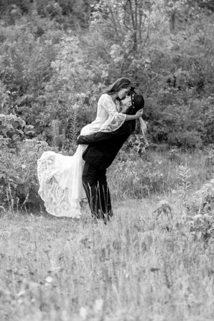 Black and white wedding photo of groom dipping bride in a meadow, bride in flowing lace dress holding bouquet.
