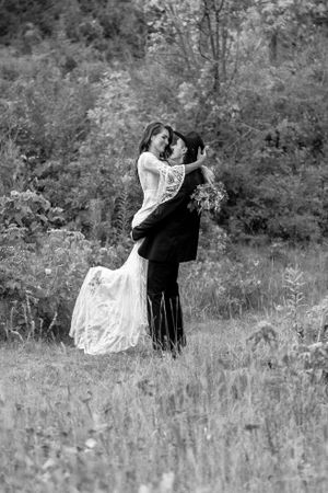 Black and white photo of bride and groom embracing and kissing in a wildflower field with lush trees behind them.