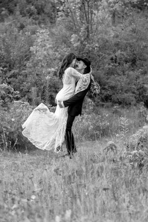 Black and white photo of groom lifting bride joyfully in a meadow, bride in lace dress with bouquet, trees behind.
