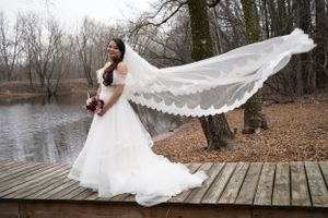 Bride in white gown with flowing lace veil blowing in wind on wooden dock by lake in autumn forest.