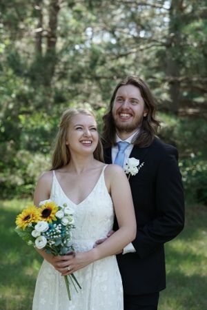 Smiling bride holding sunflower bouquet with long-haired groom in black suit in green forest.