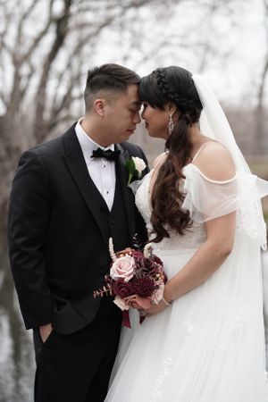 Bride and groom touching foreheads romantically, holding burgundy and pink bouquet by winter lake.