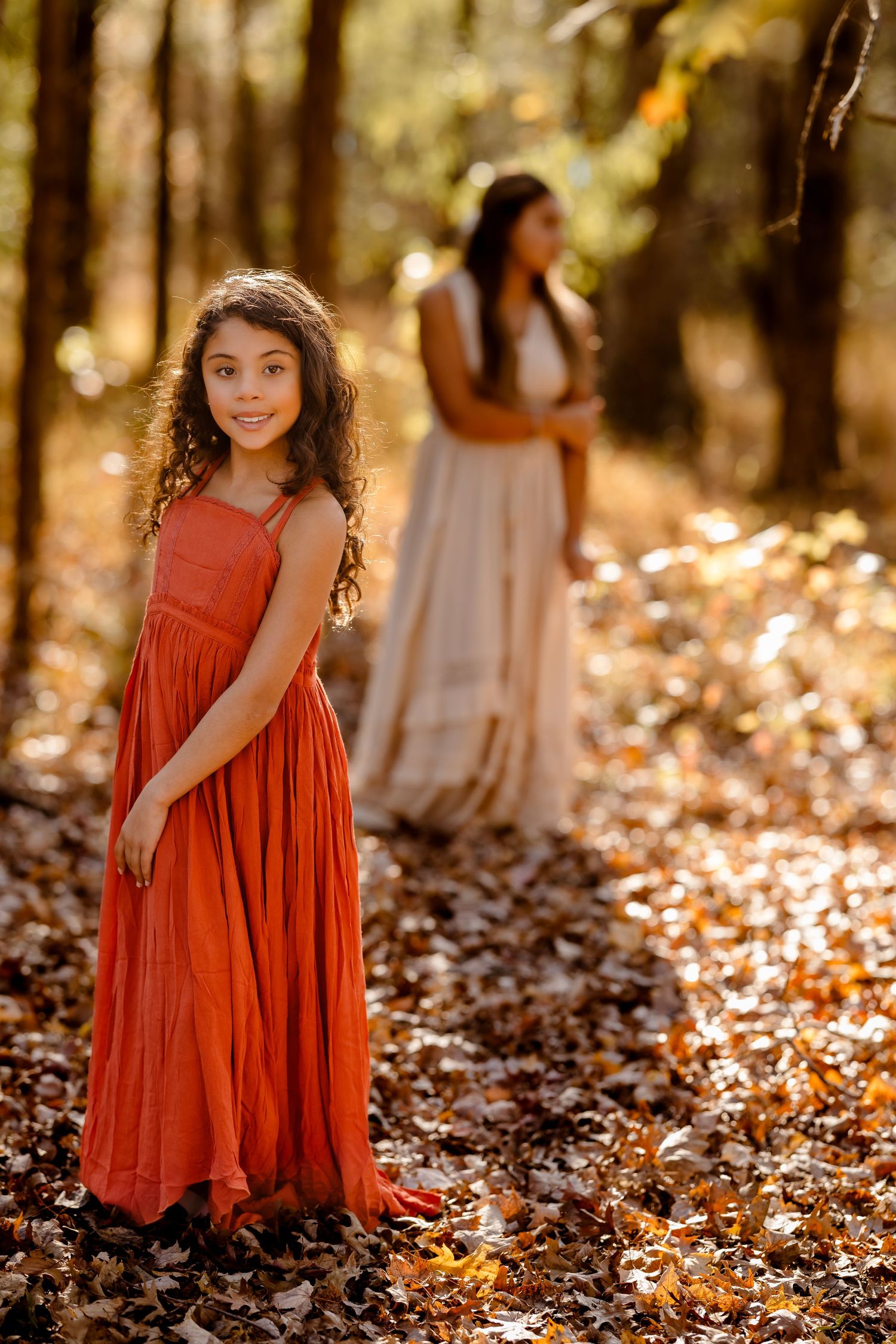 Girl in rust-colored dress among fall leaves in golden forest light.