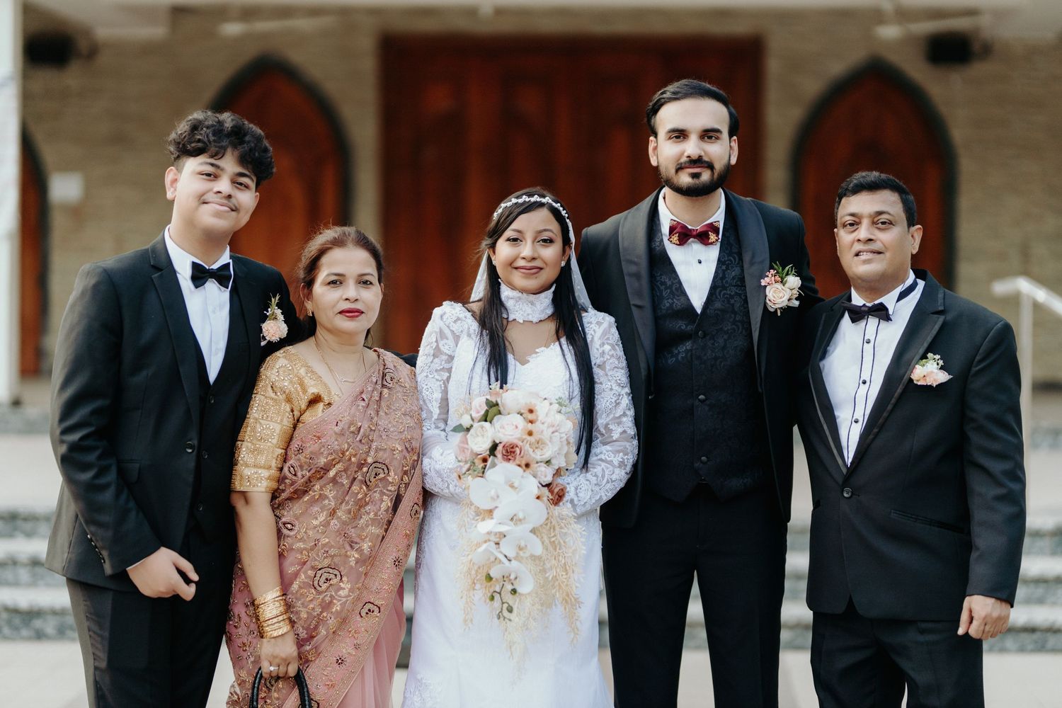 Wedding party poses in formal attire with traditional Indian sari and Western suits outside a church entrance.