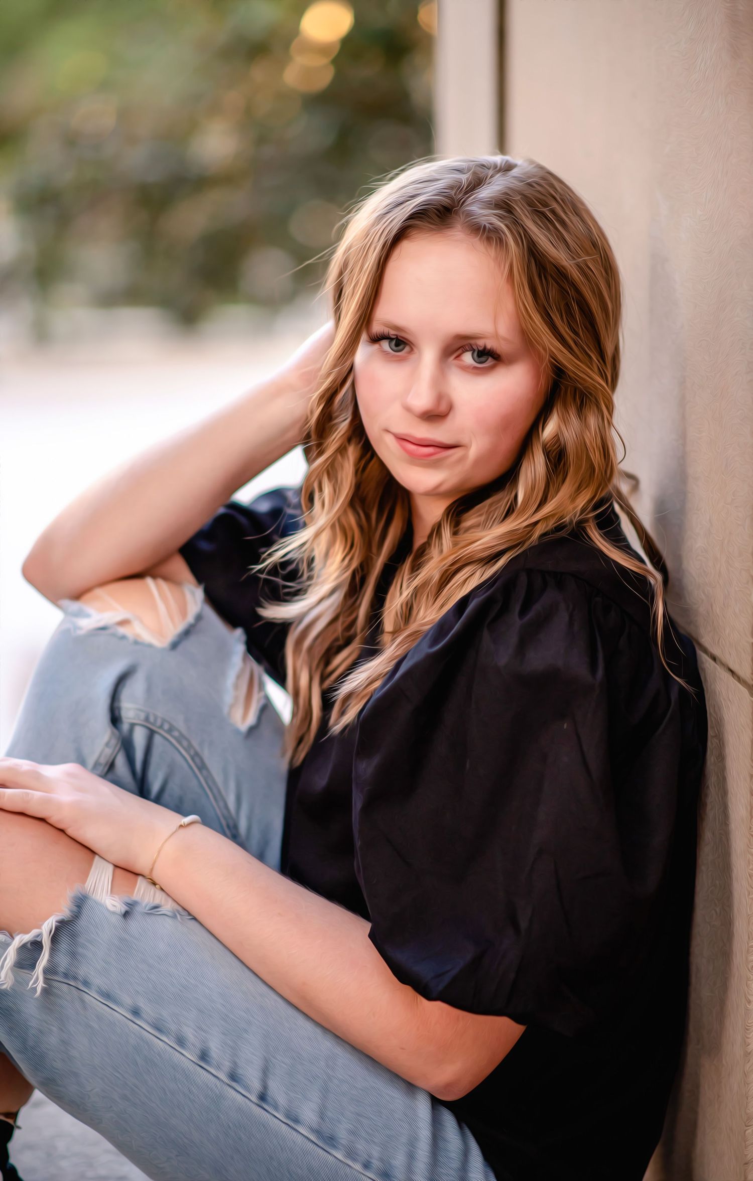 Portrait of young woman in black top and jeans sitting against wall outdoors.