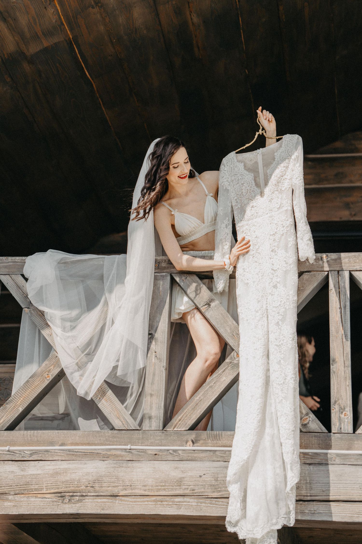 Bride in lingerie and veil admires her long-sleeve lace wedding dress hanging on a rustic wooden balcony.