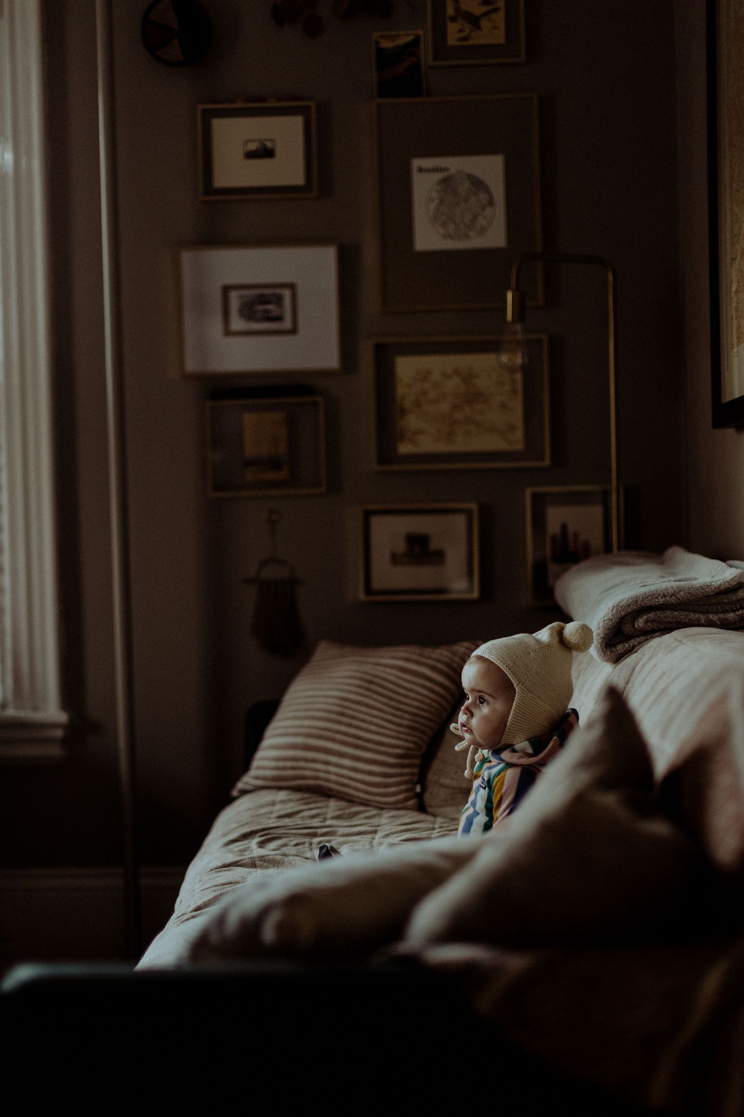 Adorable baby wearing a knit bonnet resting on a cozy sofa surrounded by framed wall art in a dim room.