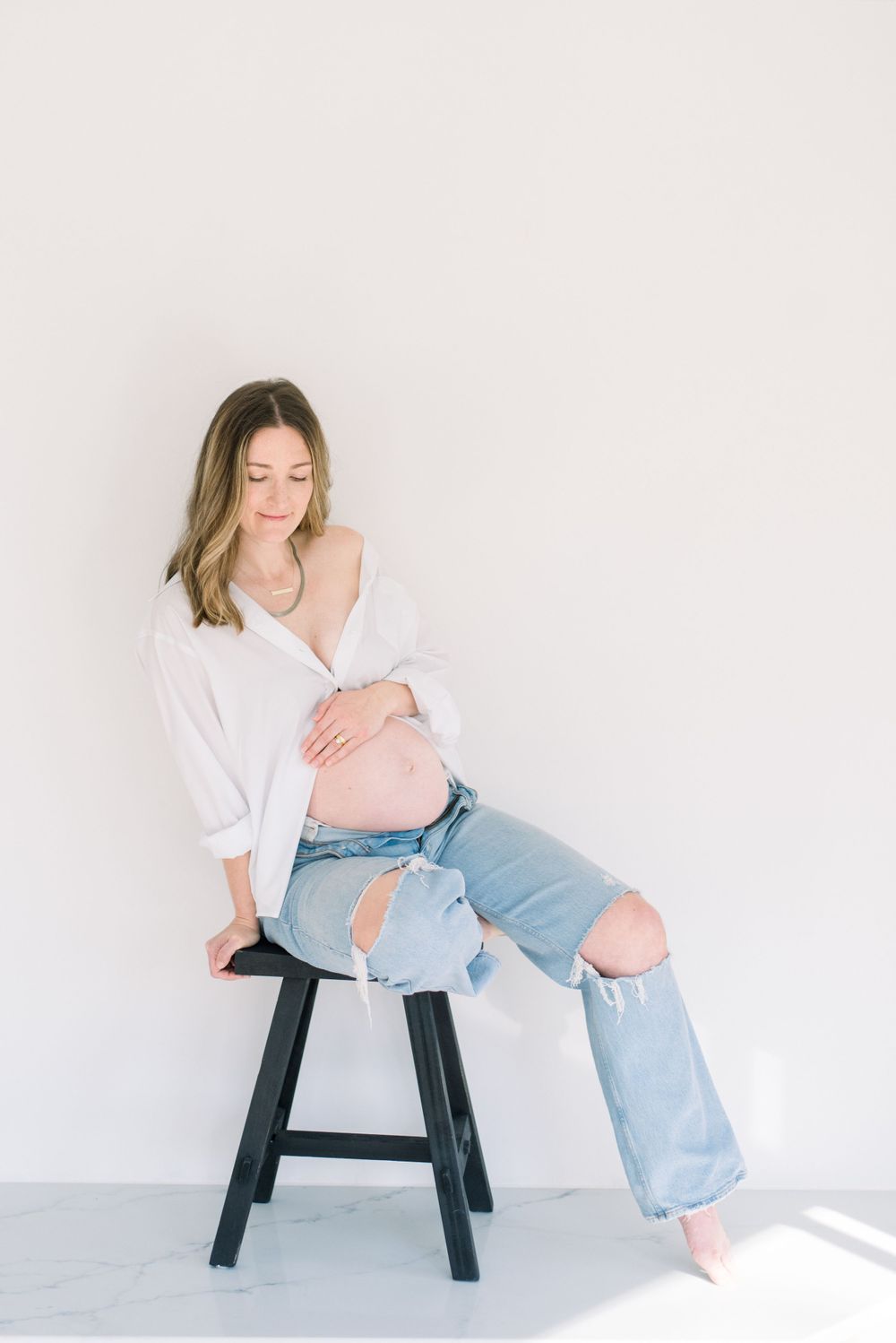 Expectant mother in white sweater and ripped jeans sits on black stool against white wall.