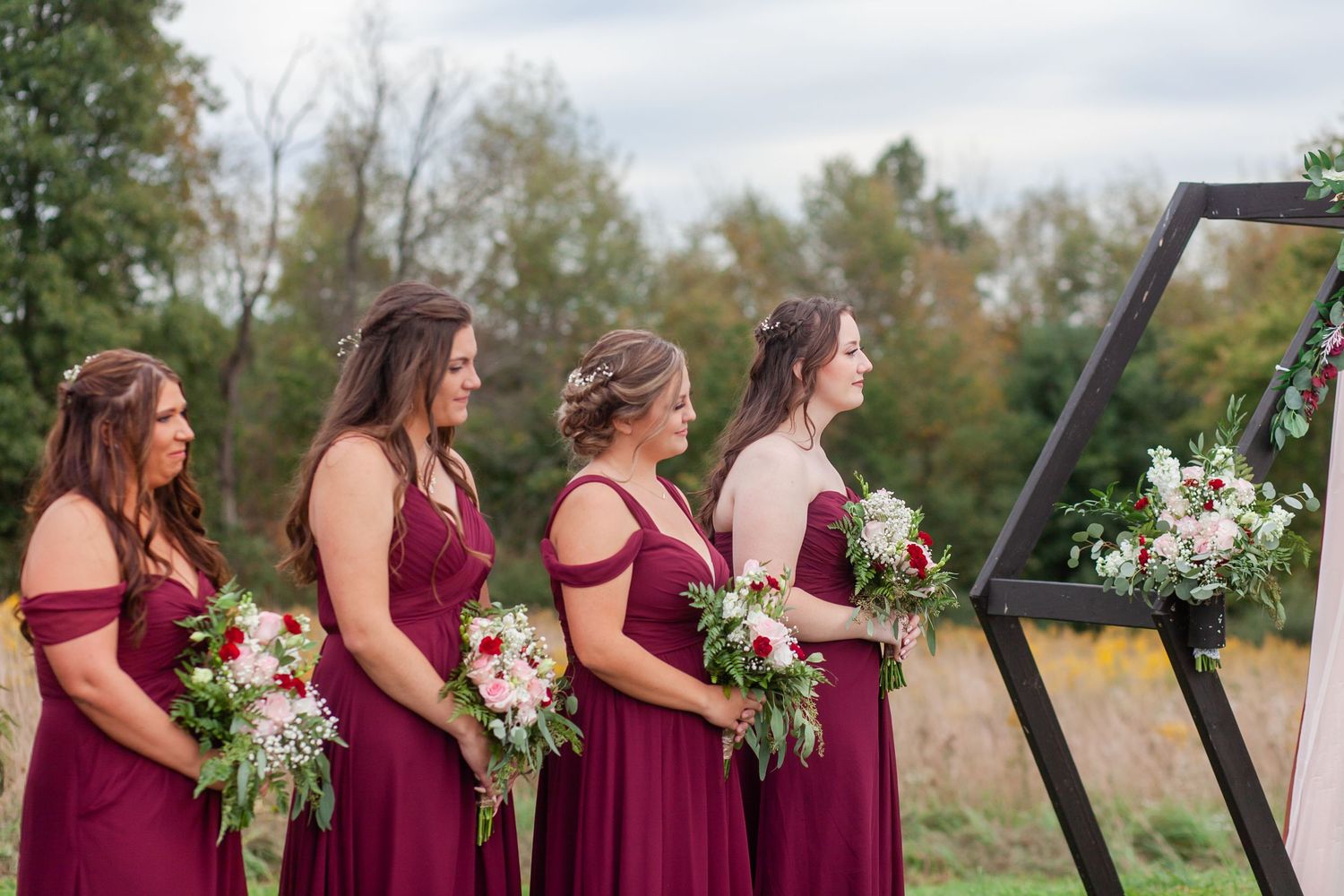 Bridesmaids in burgundy dresses hold bouquets while standing at geometric wedding arch.