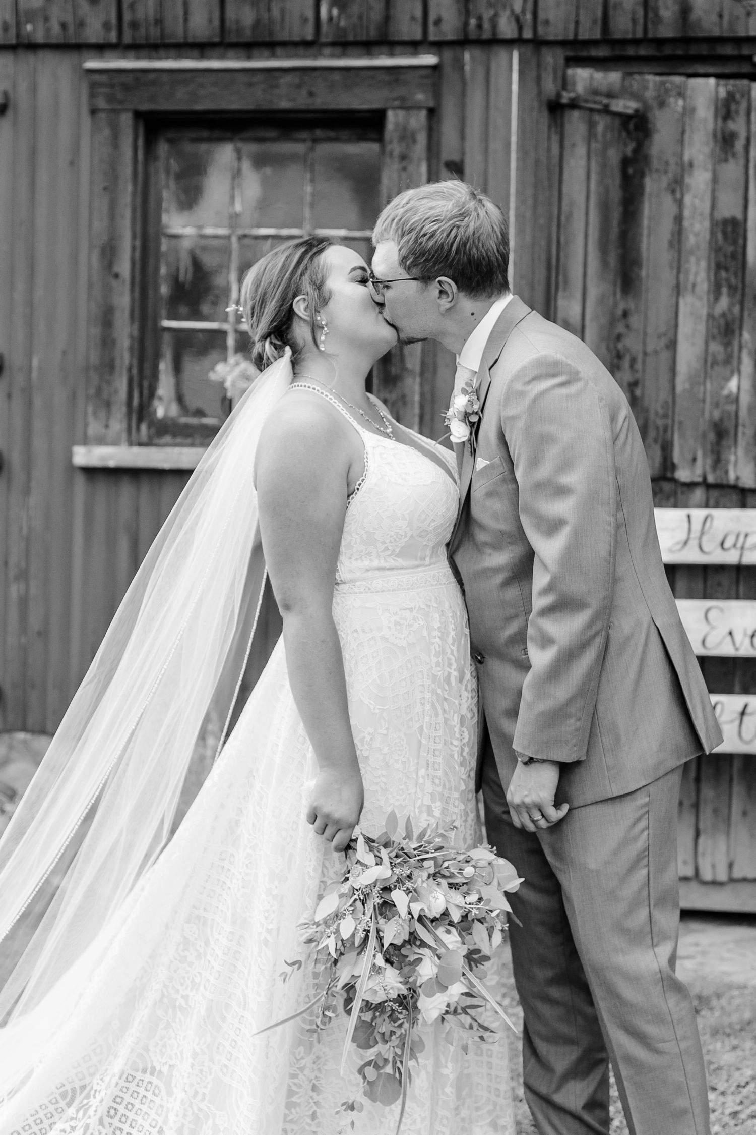 Black and white wedding portrait against rustic wooden barn backdrop.