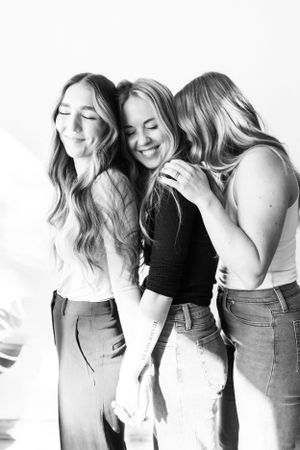 Three smiling women with long hair embracing in bright black and white portrait photo.