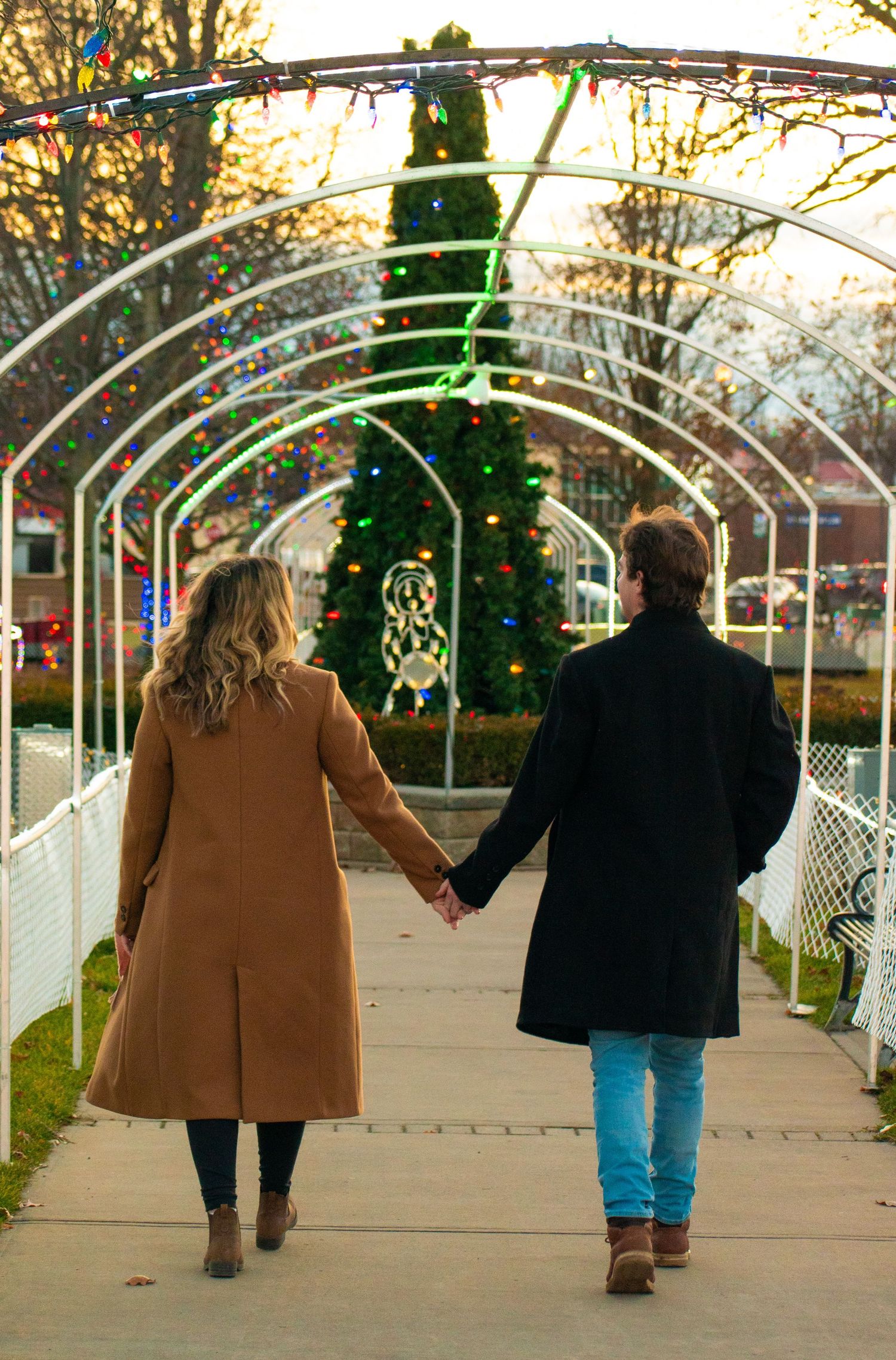 A couple holds hands while walking through a holiday light tunnel at dusk.