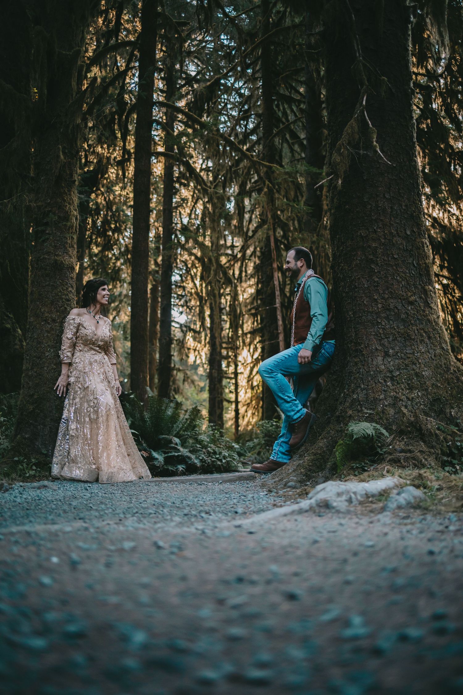 A Man and woman in wedding attire both leaning up against separate trees along a forest path the Hoh Rainforest Olympic National Park
