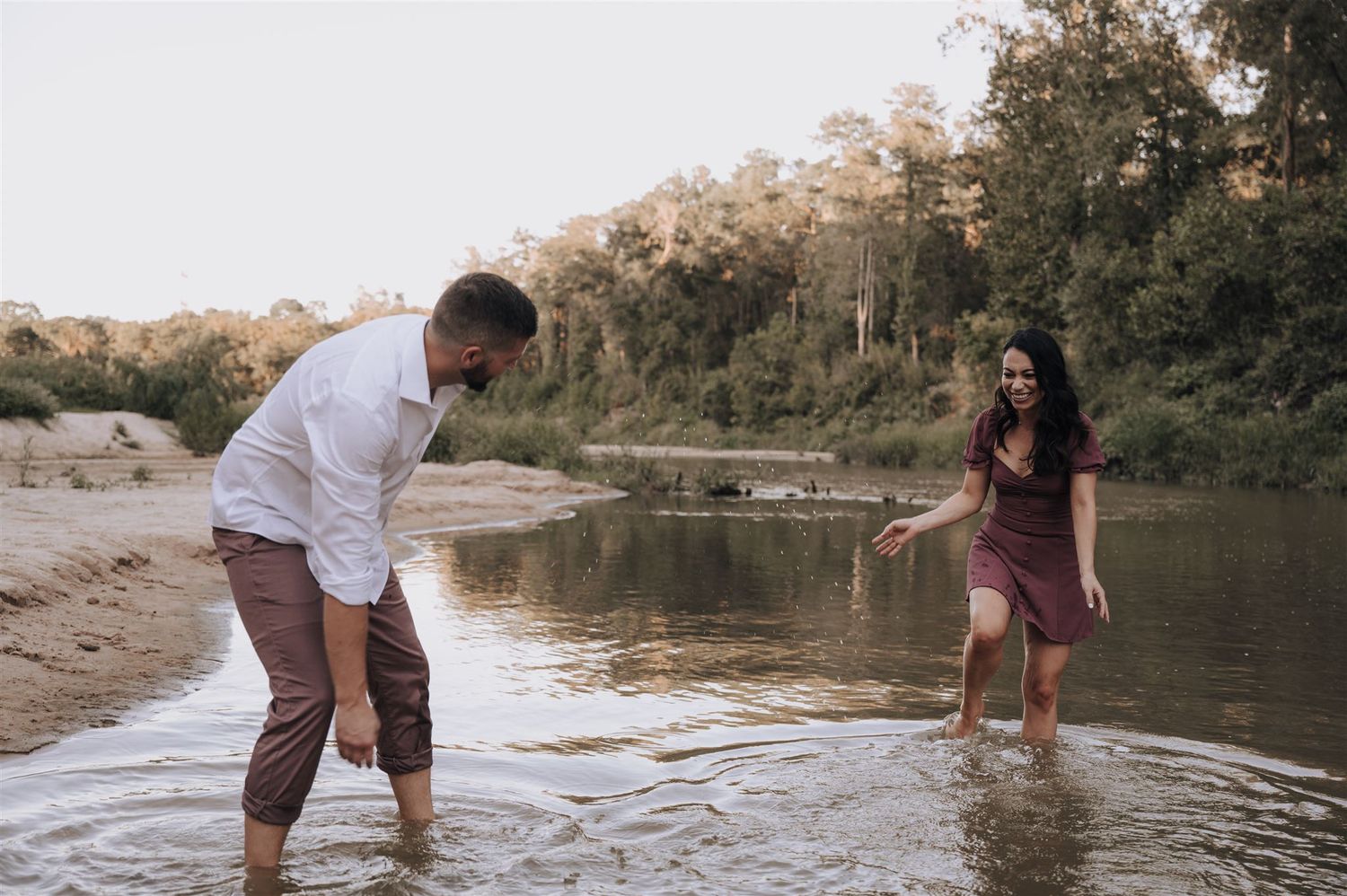 Playful couple splashing in river water wearing burgundy dress and white shirt at sunset.