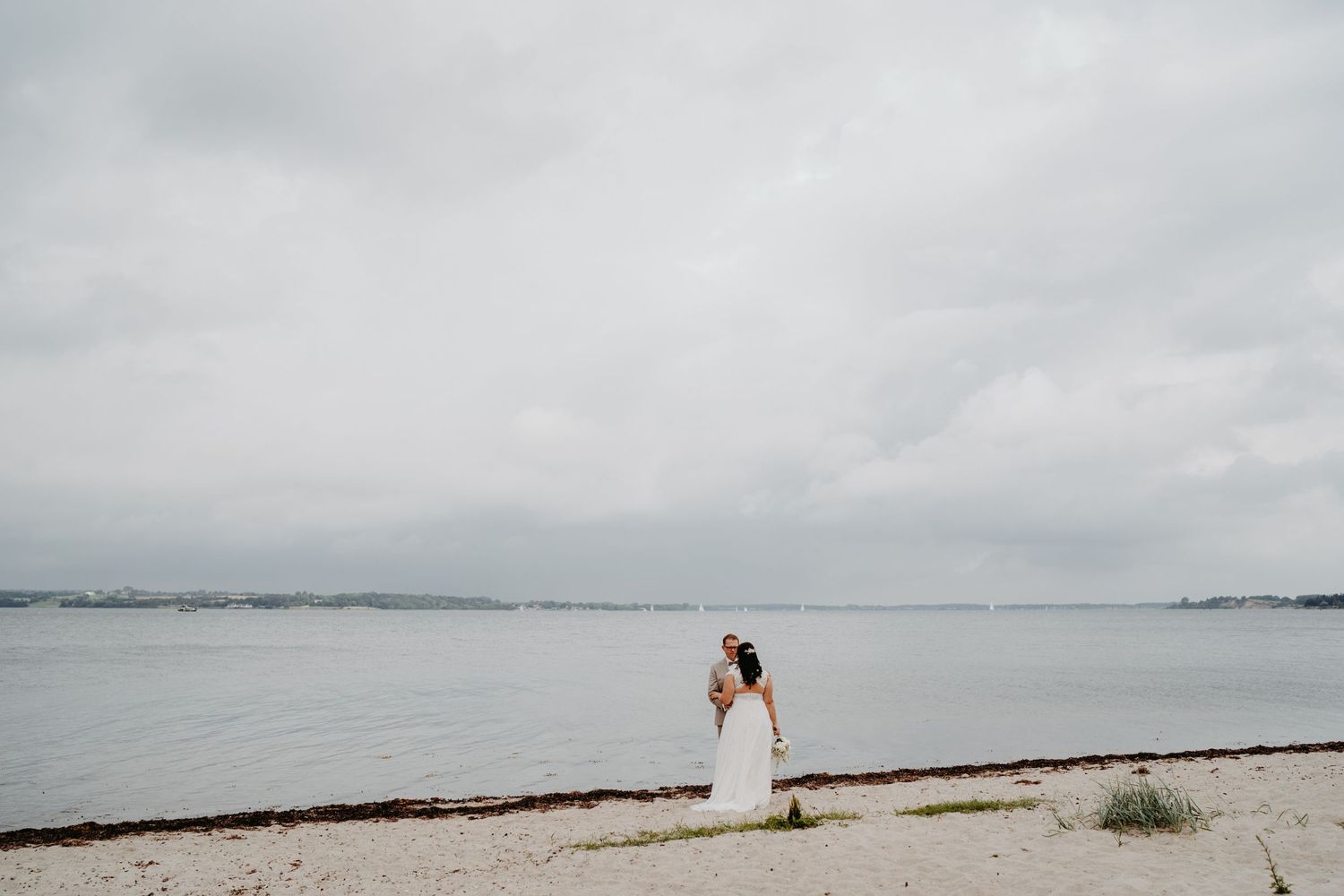 A weddingcouole in a white dress stands alone on a cloudy beach shoreline looking out over calm waters in Kiel.