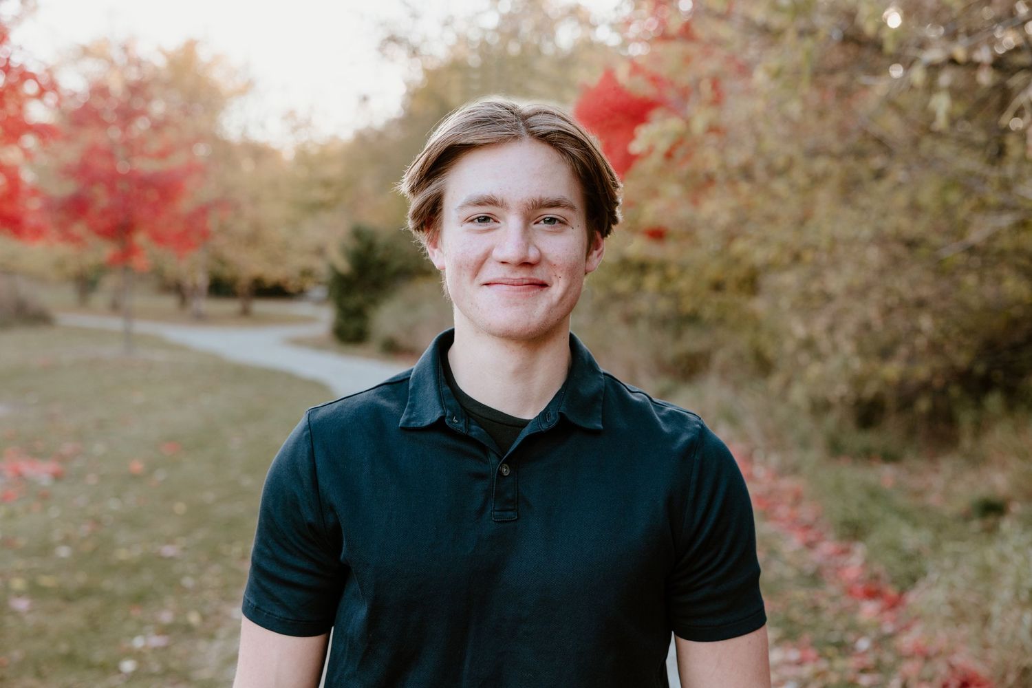 Young person in dark polo shirt standing outdoors with autumn red foliage in background.