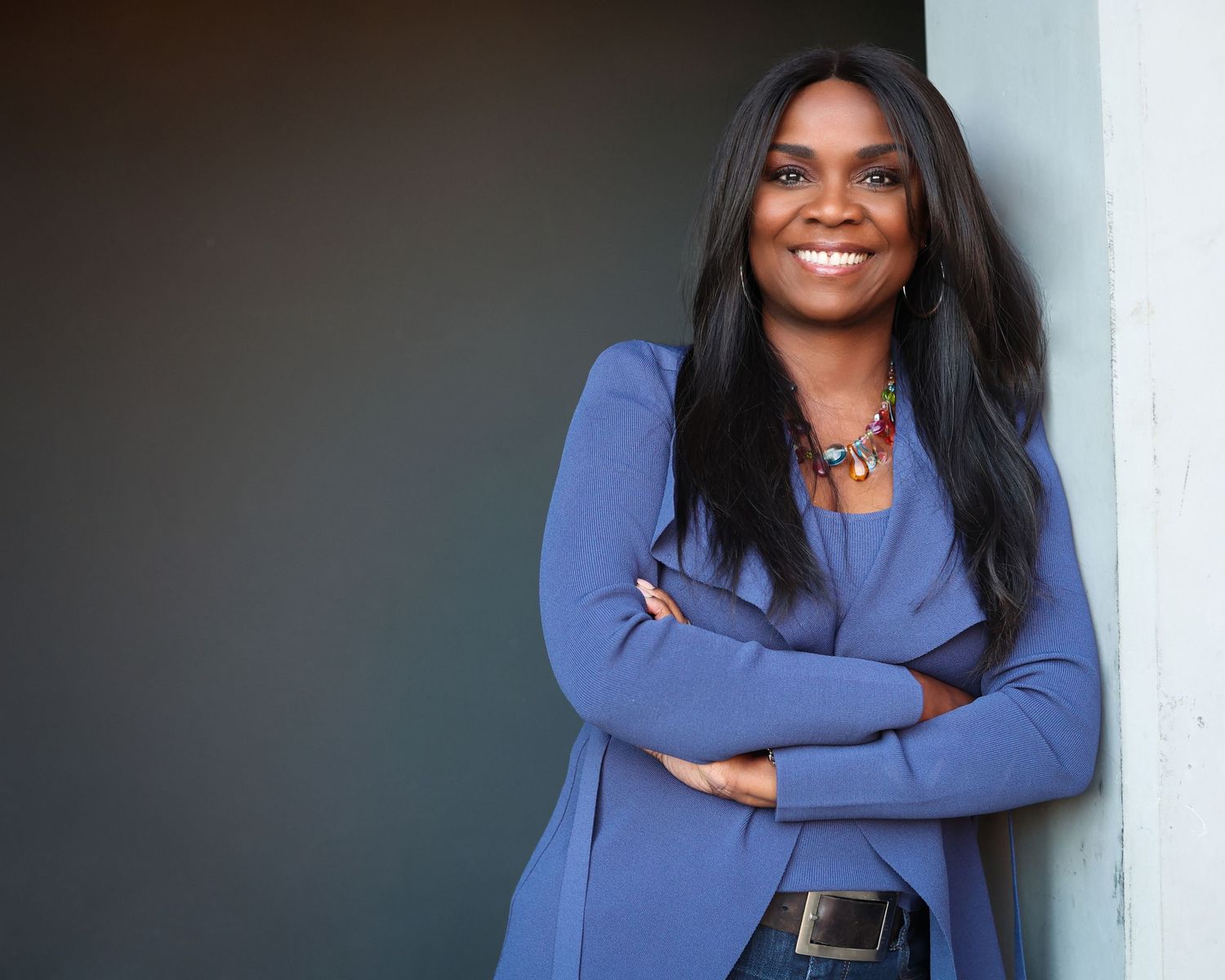 Confident Black woman in blue blazer leaning against wall with arms crossed.