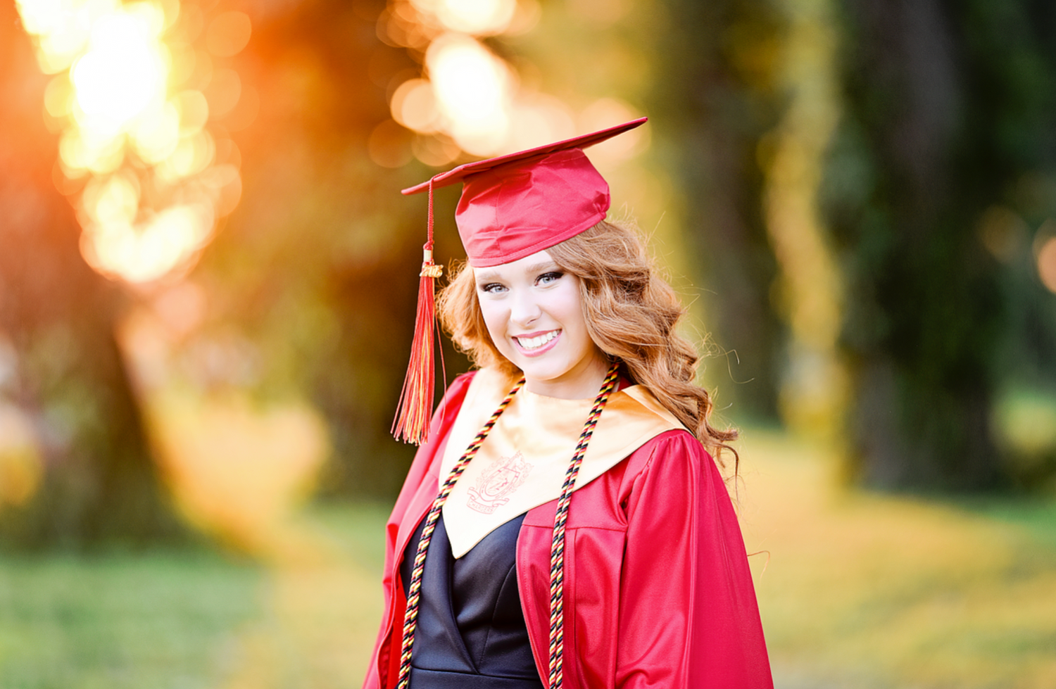 Young woman smiling in red graduation cap and gown outdoors in sunlit park setting.