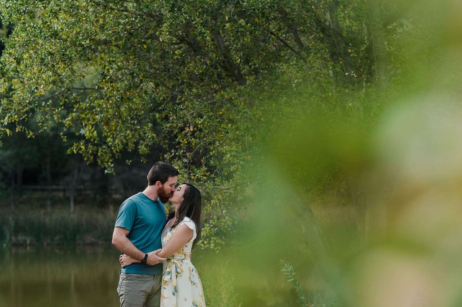 An intimate moment captured between two people by a peaceful pond with overhanging trees.
