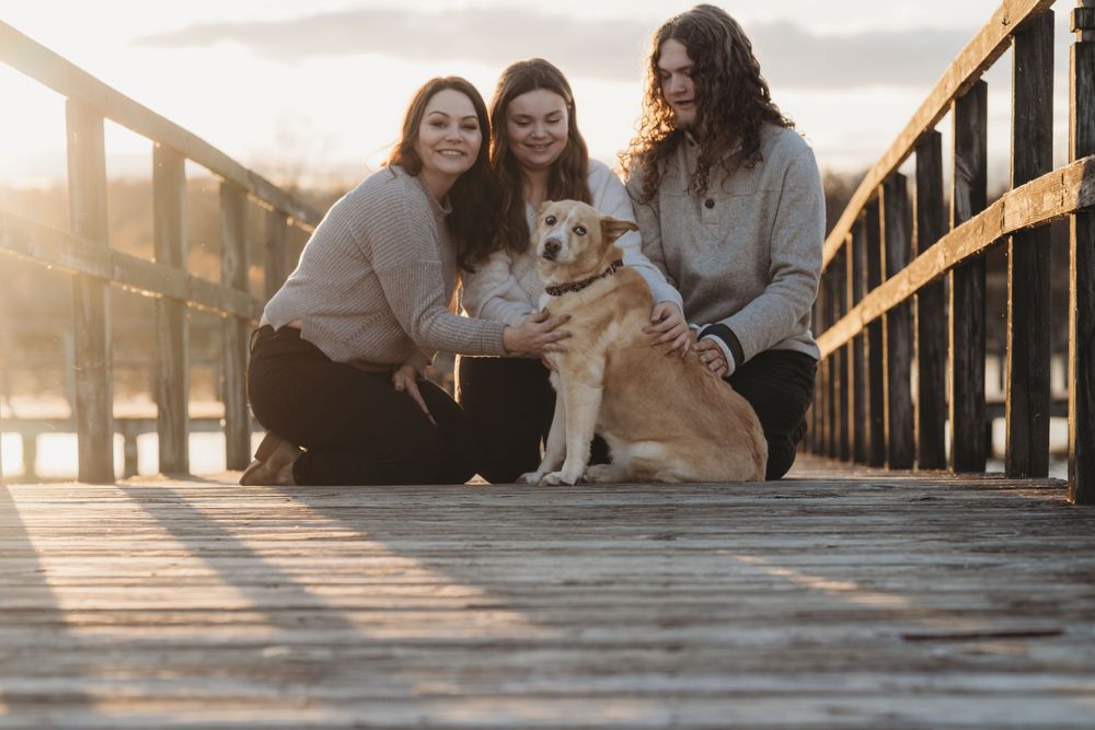 Three friends sitting with their golden retriever on a wooden dock at sunset.