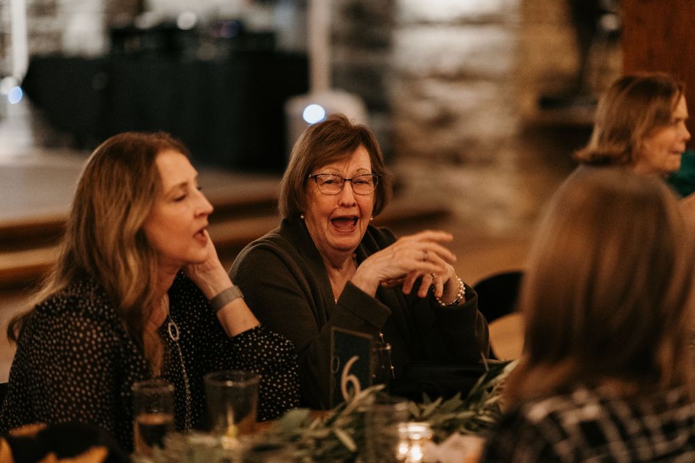 Two women laugh joyfully while seated at a decorated dinner table at a wedding reception.