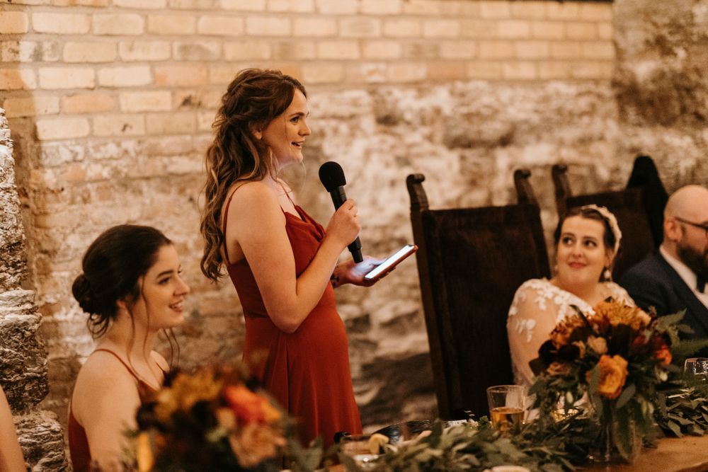 Bridesmaid in red dress delivers a heartfelt speech at a rustic stone wedding reception.
