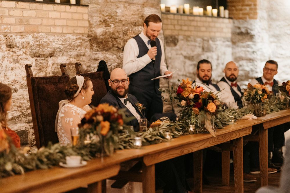 Groom in navy vest gives a heartfelt speech at a rustic wedding reception with floral centerpieces.
