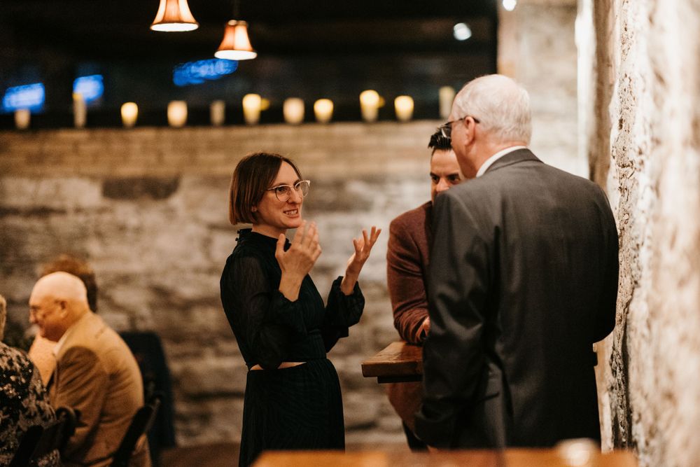 Woman in black dress gestures while conversing with a man at an elegant indoor event.