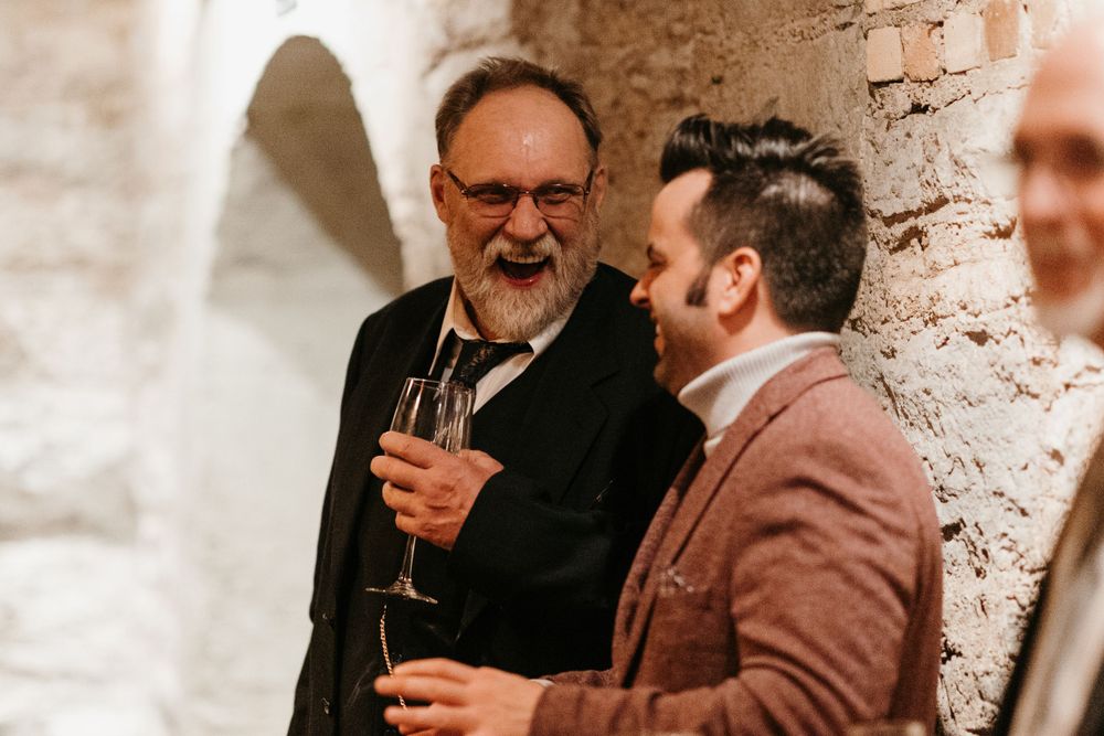 Two men smile and chat while one holds a champagne glass at a wedding reception.