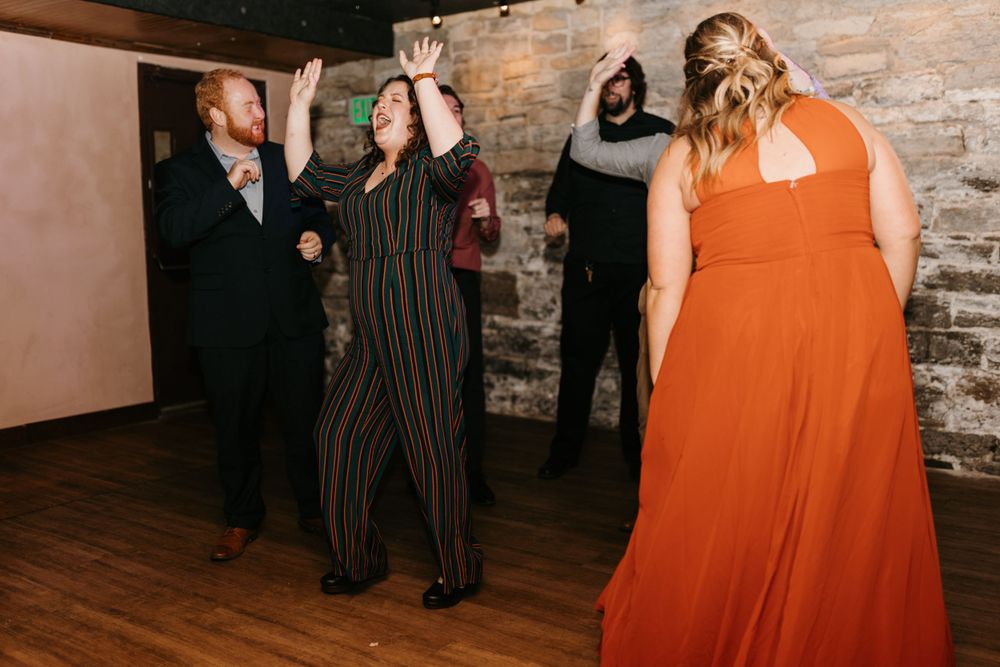 Guests in colorful outfits dance with arms raised at a lively wedding reception.