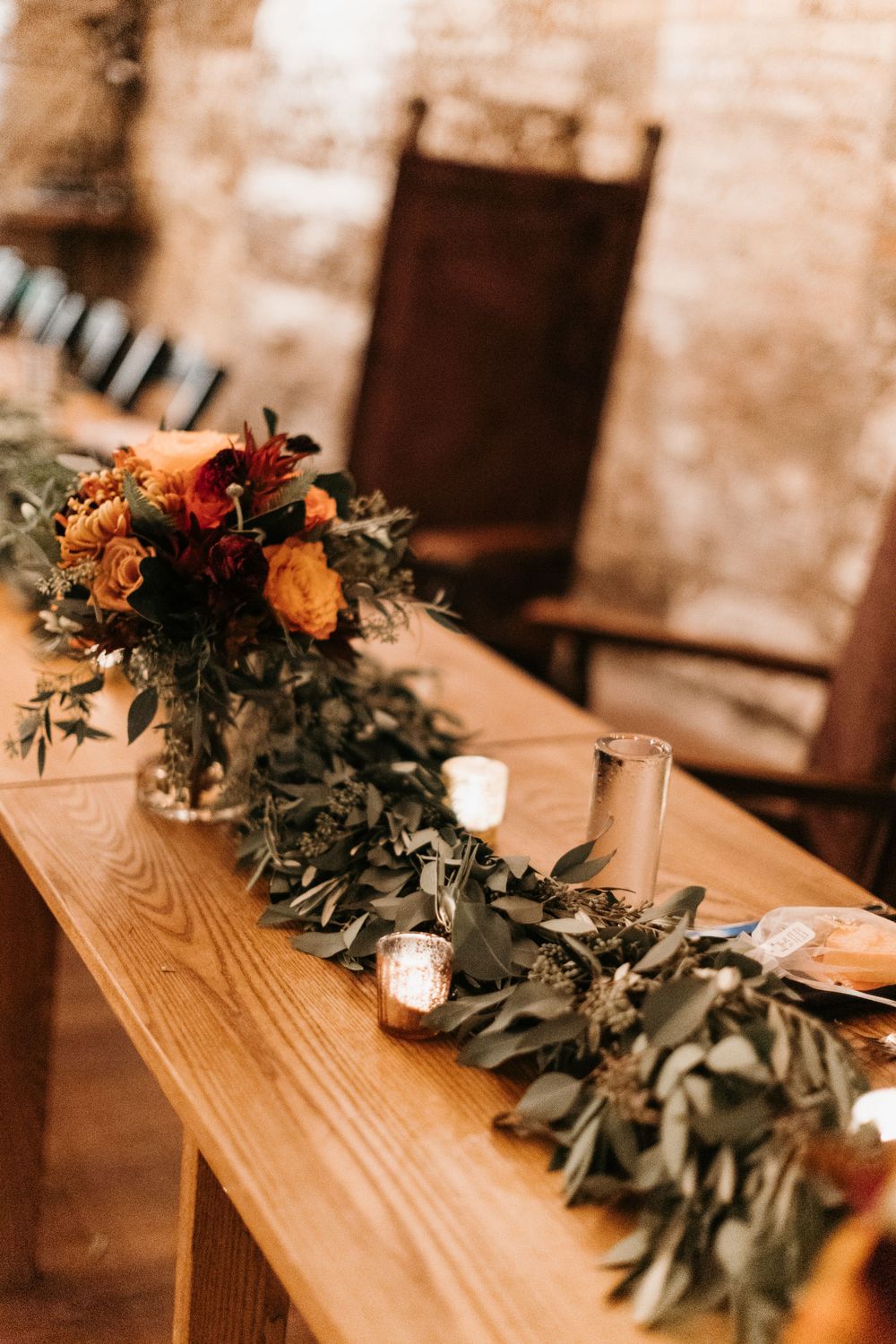 Rustic fall wedding head table with eucalyptus garland, orange floral arrangement, and glowing candles.