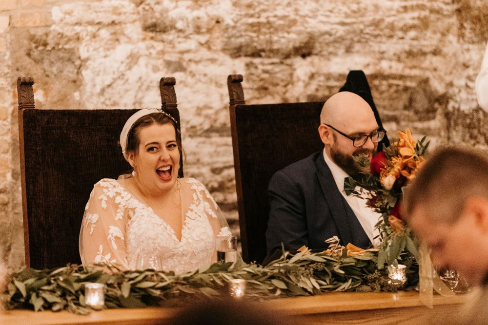 Bride laughs joyfully beside groom at sweetheart table during wedding reception speeches.