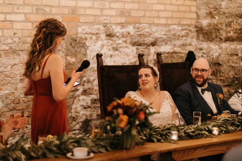 Bridesmaid in rust dress gives emotional speech as bride and groom smile at wedding reception.