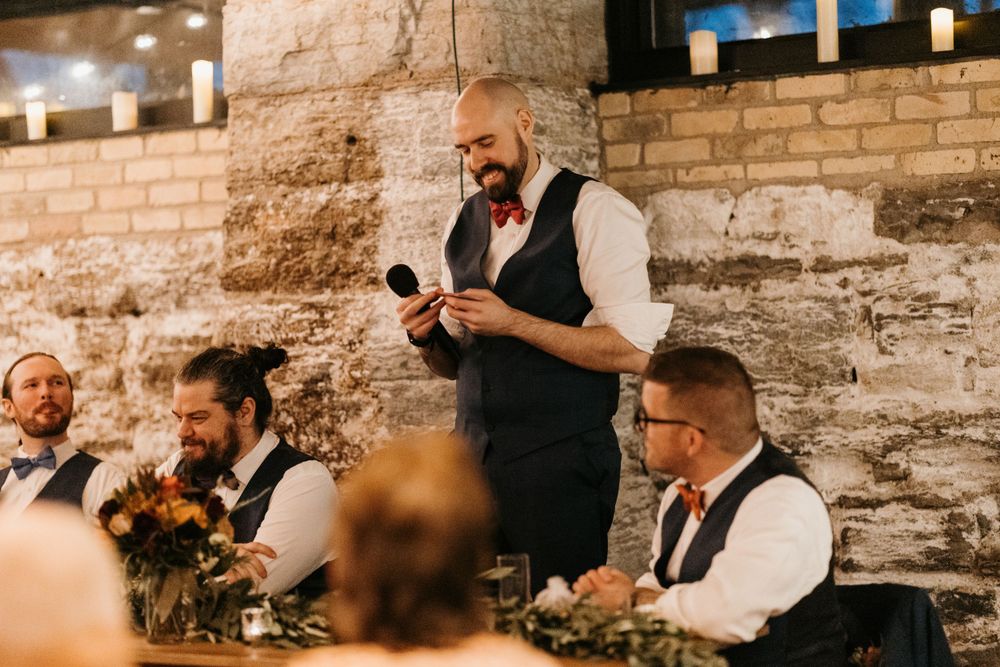 A bald groomsman in a navy vest delivers a speech holding notes at a rustic wedding reception.