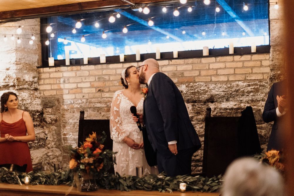 Newlyweds share romantic kiss at wedding reception in rustic brick venue with warm string lights.