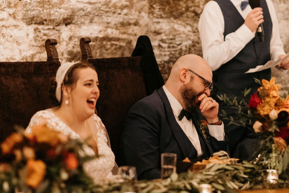 Bride and groom laugh joyfully at the head table during wedding reception speeches in a stone venue.
