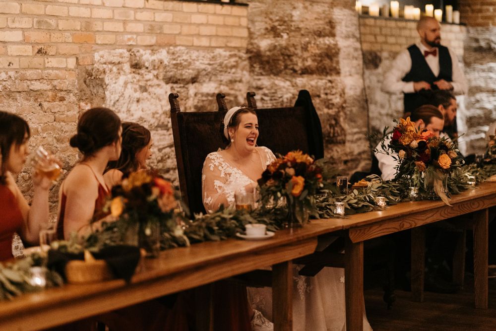 Bride laughing joyfully at head table adorned with autumn floral arrangements at wedding reception.