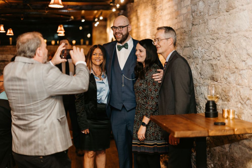 Groom in bow tie poses for a photo with family members at a rustic stone wedding reception.