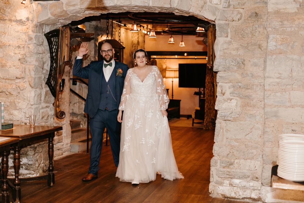 Bride and groom make grand entrance through stone archway at rustic wedding venue, groom waving joyfully.