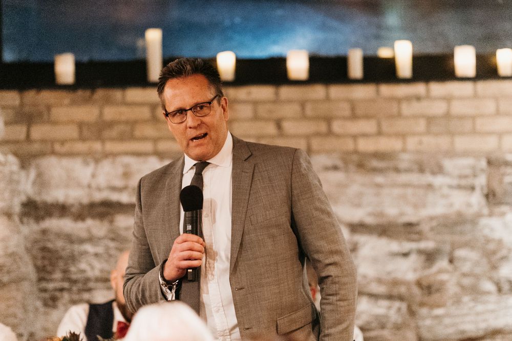 Man in gray blazer speaks passionately into a microphone at a candlelit wedding reception.