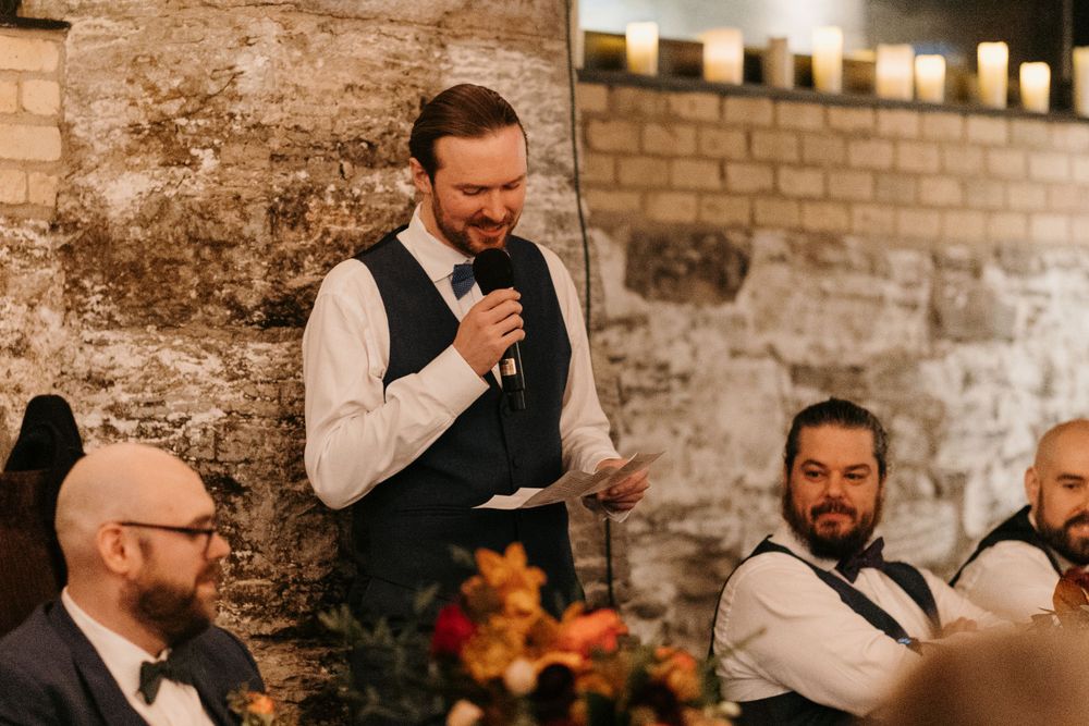 Groomsman delivers wedding toast in stone venue while guests in bow ties listen attentively.