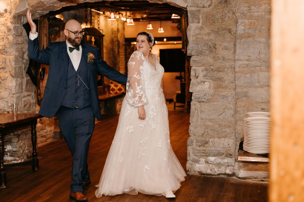 Bride in floral lace gown and groom in navy bow tie walk through stone archway at rustic wedding reception.