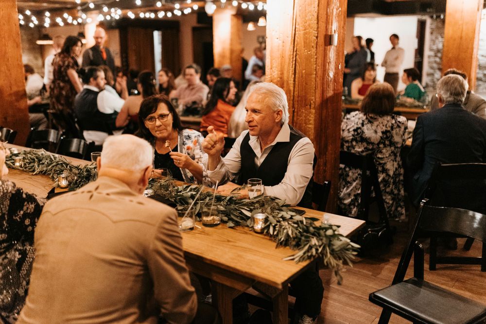 Wedding guests conversing at a rustic wooden table decorated with greenery garlands.