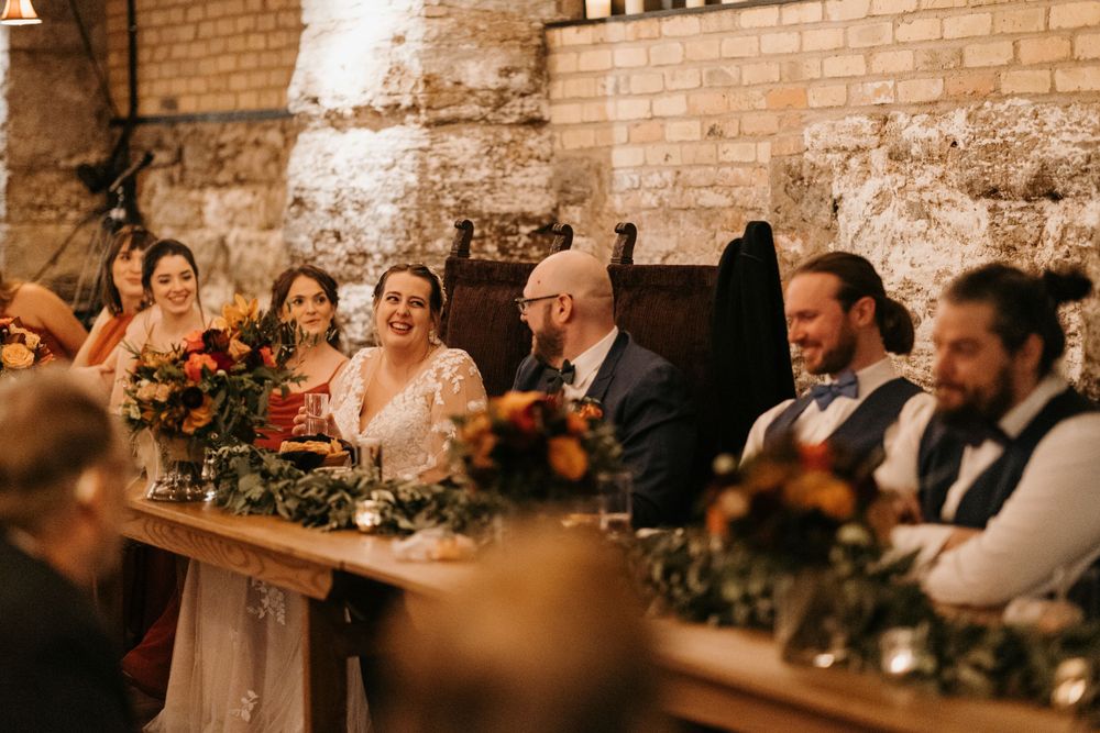 Bride and groom laugh with wedding party at a rustic stone reception hall with autumn florals.